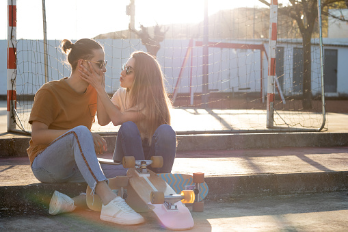 Cute young teenage couple having fun and joking with skate board around at sunset with flares around in summer time