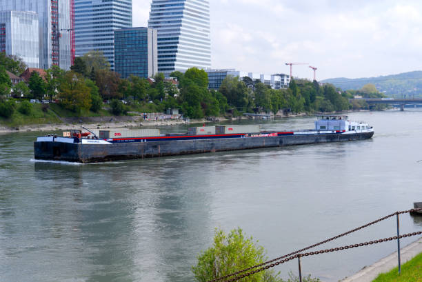 Container ship with Roche Towers at City of Basel with Rhine River in the foreground on a blue cloudy spring day. Photo taken April 27th, 2022, Basel, Switzerland.