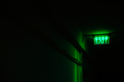 An exit sign illuminated in bright neon green stands out against the darkness of a sinister-looking corridor.