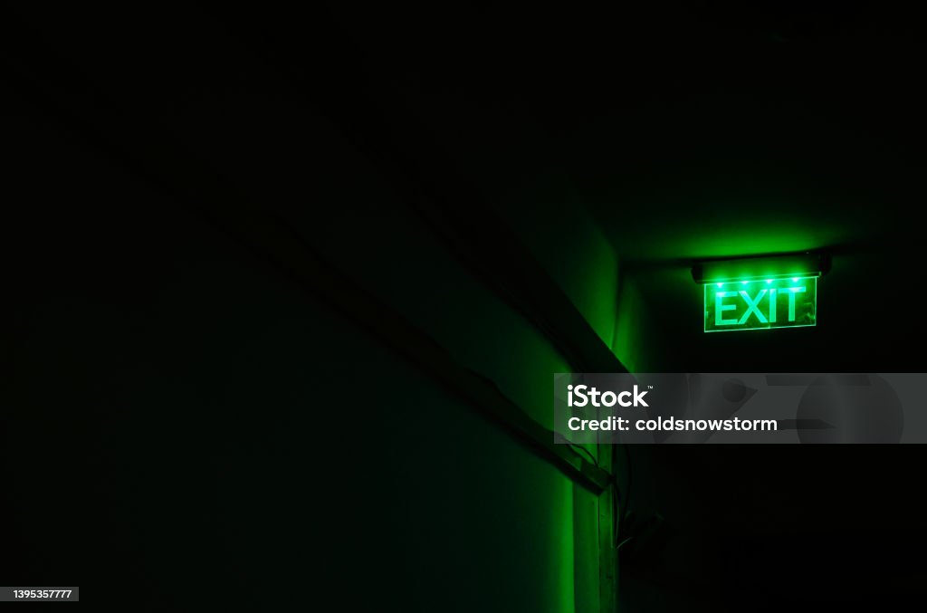 Neon green exit sign illuminated in dark corridor An exit sign illuminated in bright neon green stands out against the darkness of a sinister-looking corridor. Exit Sign Stock Photo