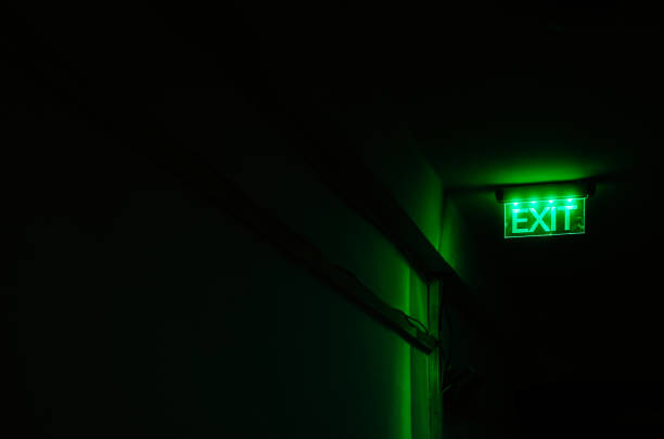 Neon green exit sign illuminated in dark corridor An exit sign illuminated in bright neon green stands out against the darkness of a sinister-looking corridor. exit sign photos stock pictures, royalty-free photos & images