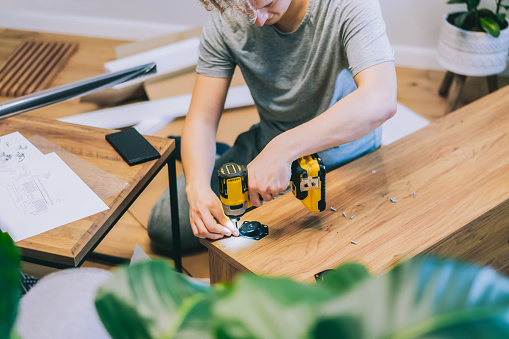 A young man with an electrical screwdriver assembles a tv stand console according to instructions in his new house. Man assembling furniture at home using a cordless screwdriver. Selective focus