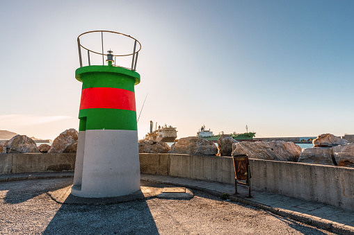 Small green, red and white coastal lighthouse with the sun on its back with large ships in the background, Motril, Andalusia Spain