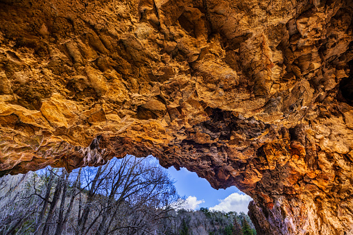 View Oit Cave Entrance - Wide angle view of limestone rock cave looking out into trees and blue sky.