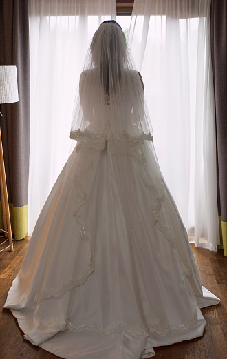 portrait of caucasian beautiful attractive woman bride in traditional european white dress standing by the window and opening curtains