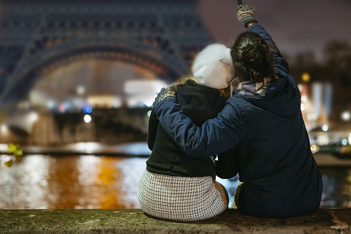 Close-up of Couple, a woman with blond hair, wearing a black jacket, a white hat and skirt, a Man with black hair tied in a braid, wearing a blue jacket, sitting next to each other on a concrete wall at seine river in front of Eiffel Tower, Paris in the evening with lighting, Man pointing with his finger on the Tower while hugging his girlfriend, rear view, focus on forefront, horizontal