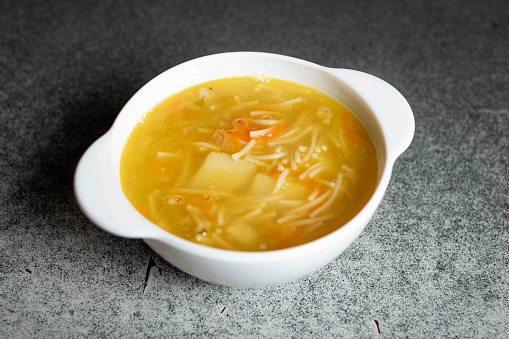Chicken soup with noodles, potatoes, onions and carrots in a white deep plate on a gray background.
