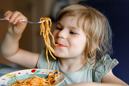 Adorable toddler girl eat pasta spaghetti with tomato bolognese with minced meat. Happy preschool child eating fresh cooked healthy meal with noodles and vegetables at home, indoors