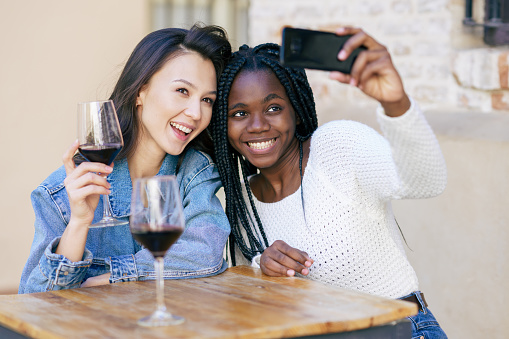 Two friends making a selfie sitting at a table outside a bar while drinking a glass of red wine.. Multiethnic women.