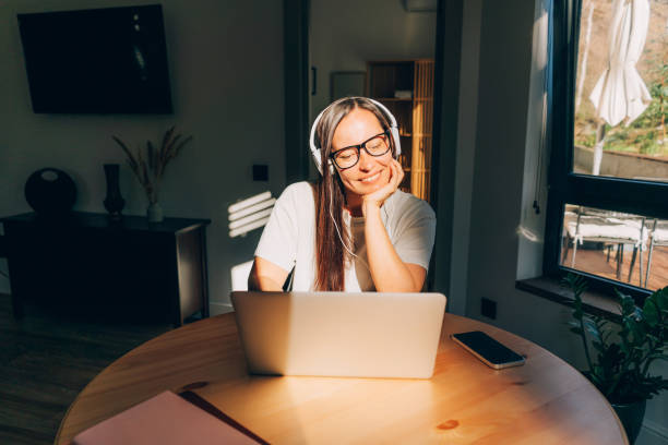 giovane donna felice che si siede al tavolo a casa alla luce del sole ascoltando l'audio nelle cuffie nel computer portatile. - mid adult audio foto e immagini stock