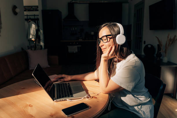la donna seduta a un tavolo a casa alla luce del sole ascolta l'audio sulle cuffie e usa un laptop. - mid adult audio foto e immagini stock