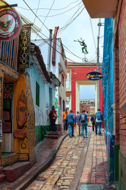 Bogotá, Colombia - Local Colombians And Tourists On The Cobblestoned Calle del Embudo, In The Historic La Candelaria District of The Andes Capital City Bogota, Colombia - July 20, 2016: Tourists and local Colombian people walk up the narrow Calle del Embudo which gets its name from its shape. The English translation of the name would be, "Funnel Street." At this end it is at its narrowest. It is obvious that only two people can walk down the street at this point at the same time. It is one of the most colorful streets in the historic La Candelaria district of Bogotá, the Andean capital city of the South American country of Colombia. Constructed over 450 years ago, when people mainly travelled on horseback, the street leads to the Chorro de Quevedo, the plaza where it is believed the Spanish Conquistador, Gonzalo Jiménez de Quesada founded the city in 1538. Ahead is the tall gate that leads into the Square. Many street facing walls in this area are painted with either street art or the legends of the pre-Colombian era, in the vibrant colours of Colombia. The altitude is 8,660 feet above mean sea level. Photo shot on and overcast morning; horizontal format. calle del embudo stock pictures, royalty-free photos & images