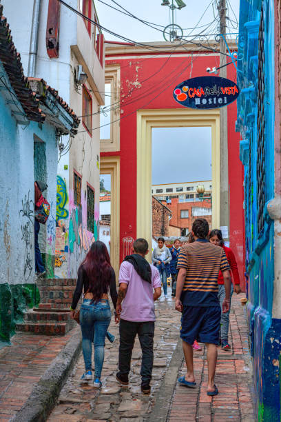 Bogotá, Colombia - Local Colombians On The Cobblestoned Calle del Embudo, In The Historic La Candelaria District of The Andes Capital City Bogota, Colombia - July 20, 2016: Local Colombian people walk up the narrow Calle del Embudo which gets its name from its shape. The English translation of the name would be, "Funnel Street." At this end it is at its narrowest. It is obvious that only two people can walk down the street at this point at the same time. It is one of the most colorful streets in the historic La Candelaria district of Bogotá, the Andean capital city of the South American country of Colombia. Constructed over 450 years ago, when people mainly travelled on horseback, the street leads to the Chorro de Quevedo, the plaza where it is believed the Spanish Conquistador, Gonzalo Jiménez de Quesada founded the city in 1538. Ahead is the tall gate that leads into the Square. Many street facing walls in this area are painted with either street art or the legends of the pre-Colombian era, in the vibrant colours of Colombia. The altitude is 8,660 feet above mean sea level. Photo shot on and overcast morning; horizontal format. calle del embudo stock pictures, royalty-free photos & images