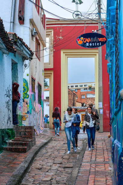 Bogotá, Colombia - Local Colombians On The Cobblestoned Calle del Embudo, In The Historic La Candelaria District of The Andes Capital City Bogota, Colombia - July 20, 2016: Local Colombian people walk up the narrow Calle del Embudo which gets its name from its shape. The English translation of the name would be, "Funnel Street." At this end it is at its narrowest. It is obvious that only two people can walk down the street at this point at the same time. It is one of the most colorful streets in the historic La Candelaria district of Bogotá, the Andean capital city of the South American country of Colombia. Constructed over 450 years ago, when people mainly travelled on horseback, the street leads to the Chorro de Quevedo, the plaza where it is believed the Spanish Conquistador, Gonzalo Jiménez de Quesada founded the city in 1538. Ahead is the tall gate that leads into the Square. Many street facing walls in this area are painted with either street art or the legends of the pre-Colombian era, in the vibrant colours of Colombia. The altitude is 8,660 feet above mean sea level. Photo shot on and overcast morning; horizontal format. calle del embudo stock pictures, royalty-free photos & images