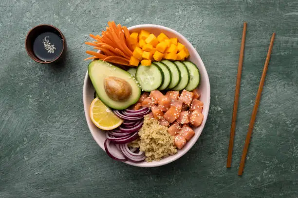 Poke bowl with salmon and avocado, quinoa, fresh cucumber, red onion, carrot and pumpkin, lemon, sesame and soy sauce for spice and to taste. Healthy food bowl.