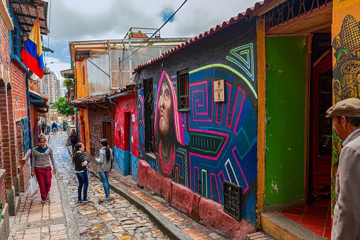 Bogotá, Colombia - July 20, 2016: A few local Colombians seen walking down the colourful, cobblestoned Calle del Embudo. The Street gets its name from its funnel shape: Embudo translates to Funnel in English. It is shaped like a funnel. It must be remembered that these streets were set up over 450 years ago when people usually travelled on horseback. The Street is well known for its Street Art, many of which feature the legends of the Pre-Colombian era. Photo shot in the late afternoon sunlight; horizontal format.