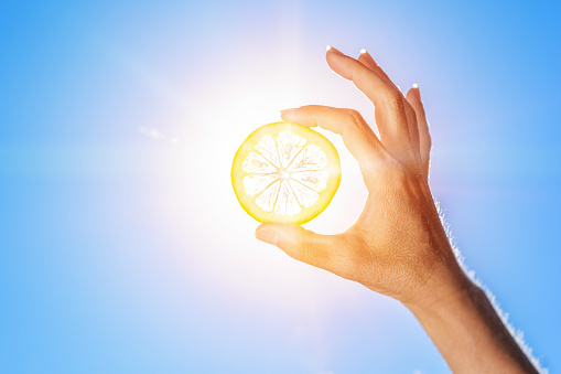 Photo of an adult female hand holding a lemon slice in front of the sun to let sunlight shine through.