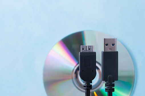 Two USB cable plugs resembling people standing in front of a CD on a blue background. Shallow depth of field.