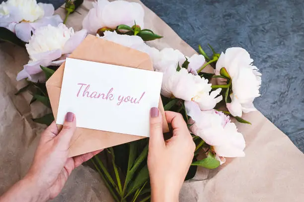 Photo of Thank you card in hands, message, envelope, pink peony flowers bouquet