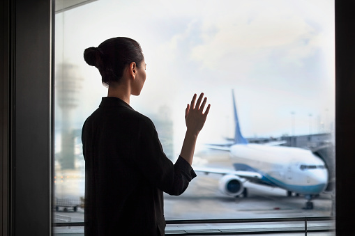 Young Chinese woman standing at airport terminal departure area waving to airplane, China