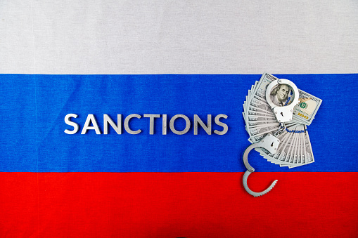 the word sanctions laid with silver metal letters on russian tricolor flag near dollar banknotes and handcuffs over it in directly above view