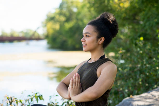 Young black woman with natural beauty is peacefully sitting in yoga meditation in prayer position by a beautiful river outdoors in the summer. She has her hair in a bun and is wearing a tank top.