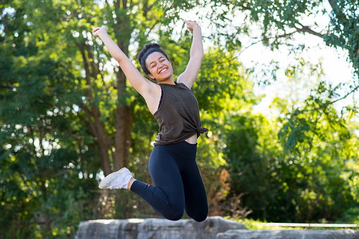 Ecstatic young black woman is smiling a toothy smile with her eyes closed as she is jumping high up with her arms in the air. She has her hair in a bun and is wearing comfortable fitness clothing.