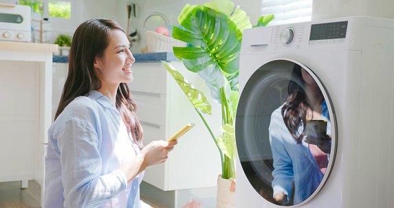 asian brunette long hair woman do the laundry in smart washing machine use smartphone operate it at home