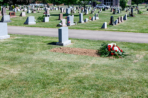 In a sunny summer cemetery, the somber flower bouquet from the recent memorial service has been placed on the dirt filled grave site. Eventually, a tombstone grave marker similar to others in the cemetery will be placed here permanently.