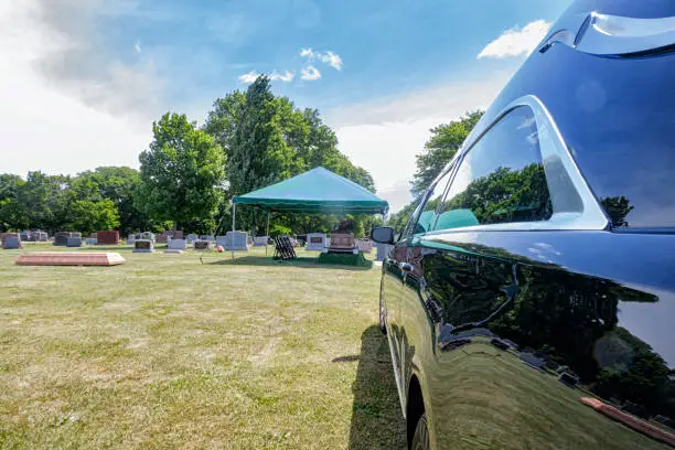 In a sunny summer cemetery, a long black funeral hearse is parked respectfully away from a small tent where a brand new casket is waiting for an imminent memorial service to begin.