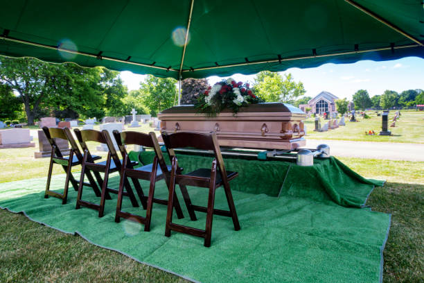 In a sunny summer cemetery, a brand new casket topped with a somber flower bouquet is waiting under a tent for a memorial funeral service to begin. Four chairs have been set up in a row next to the casket where family members of the deceased will sit during the ceremony.