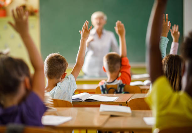 istock Rear view of elementary students raising their hands on a class at school. 1395468487