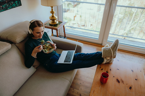 A smiling young woman using wireless headphones sitting on a couch, eating a healthy salad and streaming entertaining content on a laptop at home