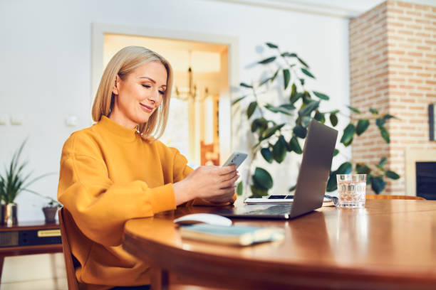 istock Smiling mid adult woman using smart phone while working remotely at home 1395474854