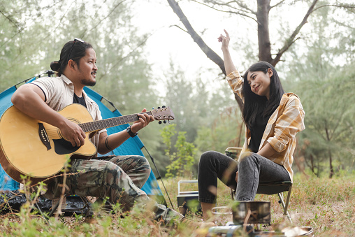 While pitching a tent in the jungle, an Asian couple plays guitar and sings.