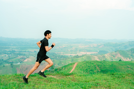 Young Asian runner, wearing black sportswear, running on a big mountain trail, cool morning, windmills, and sky in the background.