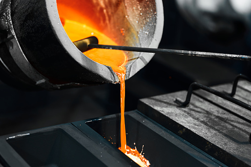 Pouring hot melt silver metal into rectangle graphite casting form from induction oven in dark plant workshop extreme closeup