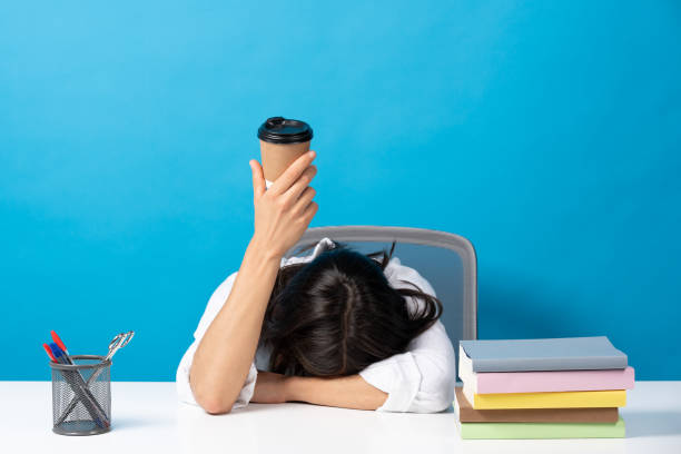 Woman head on desk showing disposable cup of coffee Woman sleeping head on desk showing disposable cup of coffee isolated on blue background. Caffeine addiction concept caffeine stock pictures, royalty-free photos & images