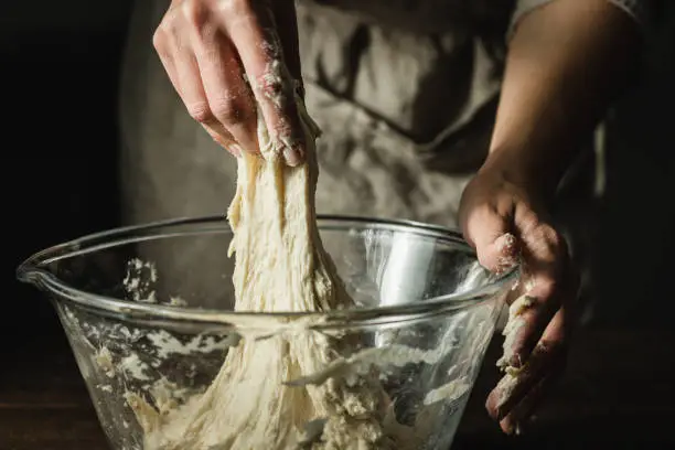 Close-up of female hands kneading dough in a glass bowl. Cropped shot of a woman preparing dough for preparing bread in kitchen.