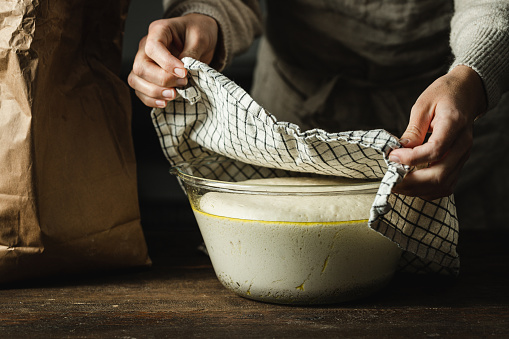 Close-up of female hands removing cover from bread dough in a large bowl after rising. Woman preparing focaccia bread in kitchen.