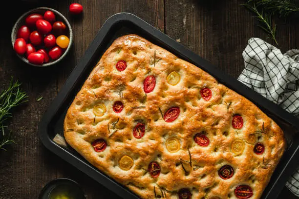 Top view of freshly baked focaccia bread with ingredients and table cloth on wooden table. Close-up of homemade focaccia bread with bowl of cherry tomatoes, rosemary and oil on table.