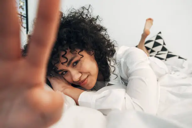 Shy young mixed race woman looking at camera lying down on the bed covering lens with hand. Selective focus on face. Lifestyle concept.