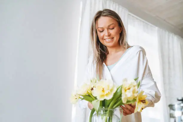 Young beautiful smiling woman forty year with blonde long hair in white shirt with bouquet of yellow flowers in hands near dinner table in bright interior at home
