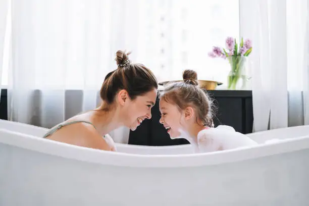 Photo of Young mother woman with long hair with little tween girl daughter in pajamas having fun in the bath with foam at home