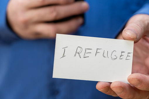 I Refugee. Inscription in jagged gray letters on paper. Man holding up two white paper rectangles with handwritten text. Close-up of the hand of a mature man.