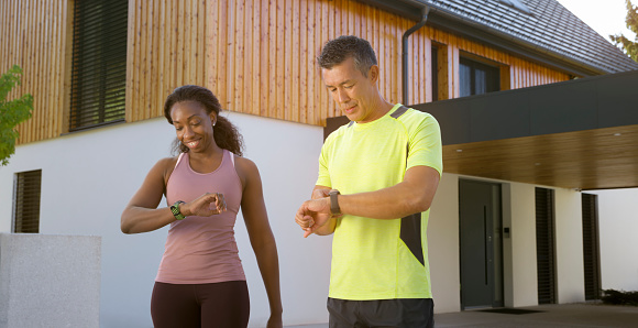 Smiling man and woman checking time on smart watch against house while standing together.