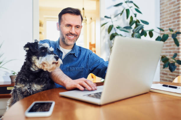 istock Cheerful man using laptop at home with dog on lap looking on a screen 1395512243