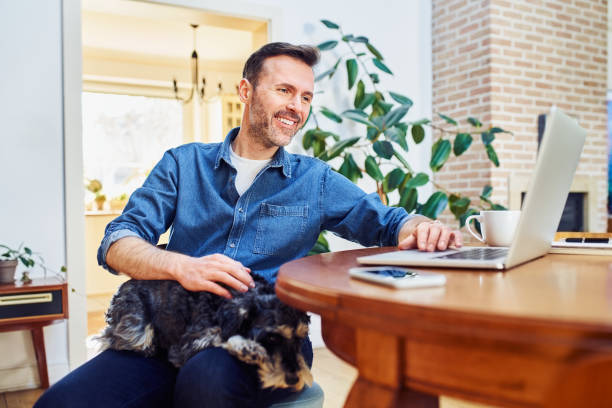istock Handsome mid adult man using laptop sitting at a table at home with dog sleeping on lap 1395512270
