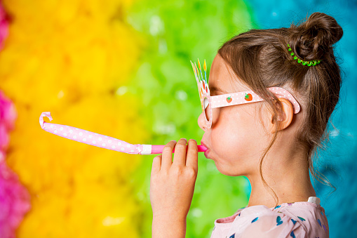 Cute girl celebrating birthday, wearing party paper glasses, blowing party horns, laughing and having fun. Bright rainbow coloured background. Happy event.