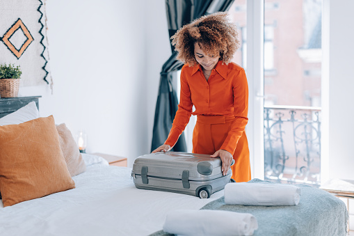 curly hair brazilian elegant young woman in orange suit open her luggage in hotel room bed